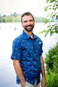 Homme dans une chemise bleue à manche courte pose devant un plan d'eau avec le sourire.